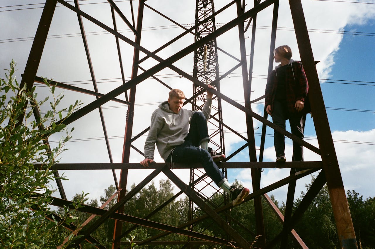 Two young adults climbing a metal transmission tower in an outdoor setting under a blue sky.